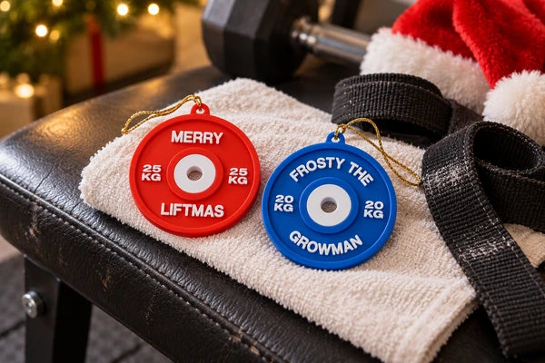 Rode Merry Liftmas- en blauwe Frosty the Growman-hanger op een handdoek naast lifting straps op een trainingsbank.